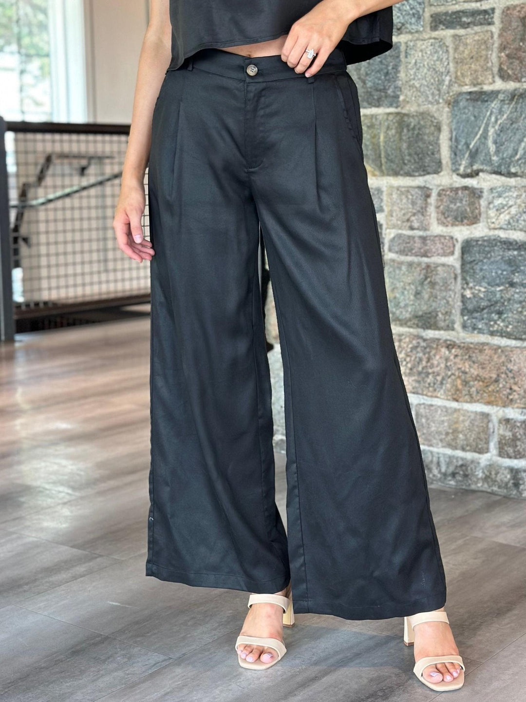 A person wearing black wide-leg cropped pants with a button closure, paired with a black crop top and beige heeled sandals, standing indoors against a stone wall and wood floor.