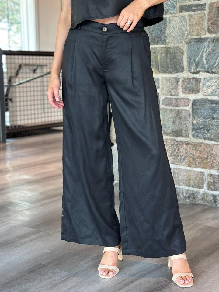 A person wearing black wide-leg cropped pants with a button closure, paired with a black crop top and beige heeled sandals, standing indoors against a stone wall and wood floor.