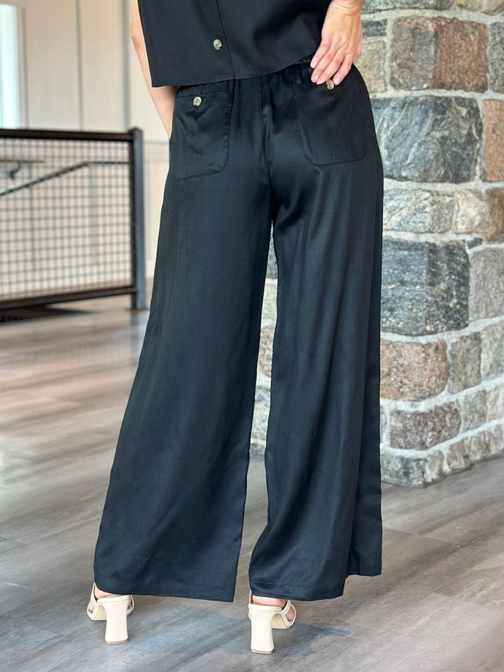 Black wide-leg pants with front patch pockets, shown on a model standing in front of a stone wall and wood floor.