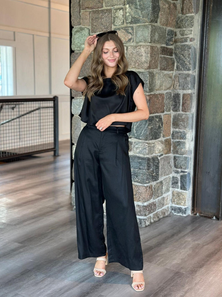 A woman modeling a black satin-like short-sleeve top and matching wide-leg trousers with a black belt, standing in an indoor setting with a stone pillar and wood floor.